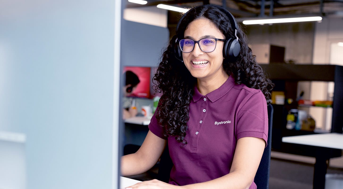A woman in a Bystronic logo purple shirt wearing headphones sits at a computer, focused on her work.
